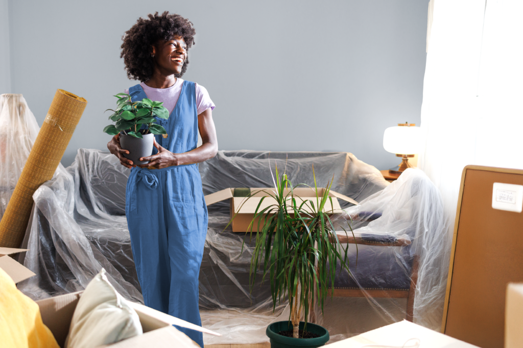 Black woman standing in living room, smiling and holding a houseplant